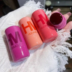 HUDA MOJI colorful lipstick collection featuring pink, coral, and red lipstick tubes with matching product packaging arranged on a soft pastel background.